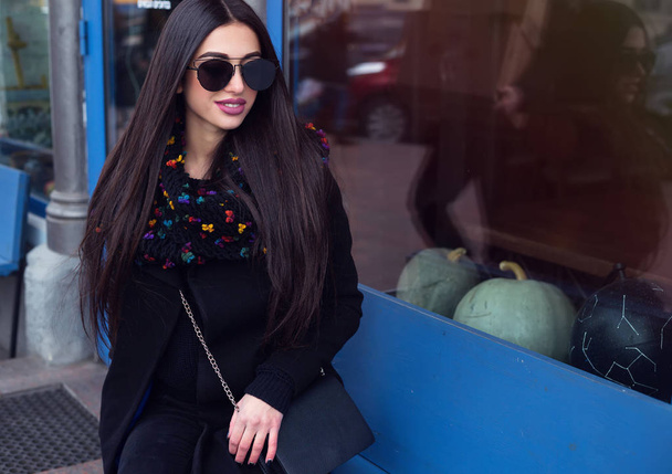 Street fashion concept - portrait of a pretty girl,sitting in the street cafe outside, Wearing hat and vintage black jacket,blue scarf, trendy sunglasses.Soft warm vintage colour tone.Copy space - Фото, зображення