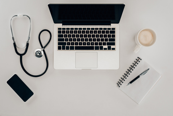 pohled shora uspořádány stetoskop, laptop, zápisník a šálek kávy na pracovišti lékaři - Fotografie, Obrázek