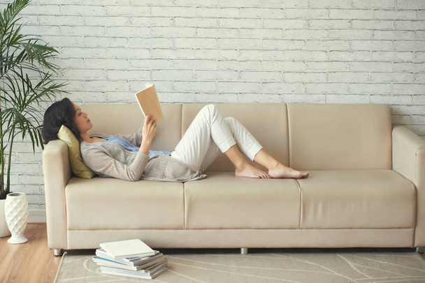 Nuori nainen makaa sohvalla ja lukee, pino kirjoja on lattialla hänen vieressään
 - Valokuva, kuva
