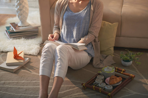 zugeschnittenes Bild einer Frau auf dem Boden sitzend mit interessantem Buch und Tablett mit Tee - Foto, Bild