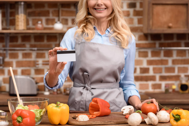 imagen recortada de la mujer mostrando tarjeta de crédito y de pie cerca de la mesa con verduras en la cocina
 - Foto, Imagen