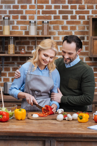 husband hugging wife while she cutting vegetables in kitchen  - Fotó, kép