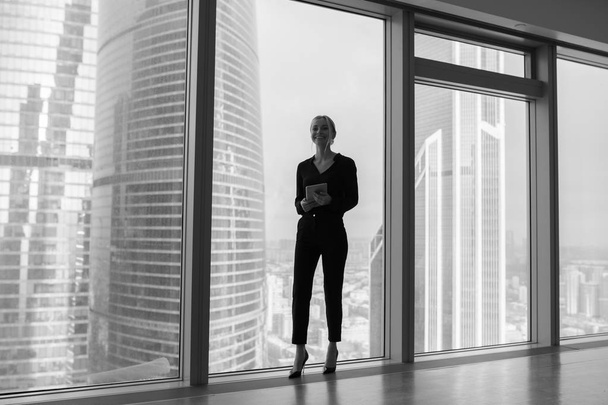 Biznes kobieta trzymając tablet i stojąc w nowoczesnym biurze. Panoramiczne okna tło. - Zdjęcie, obraz