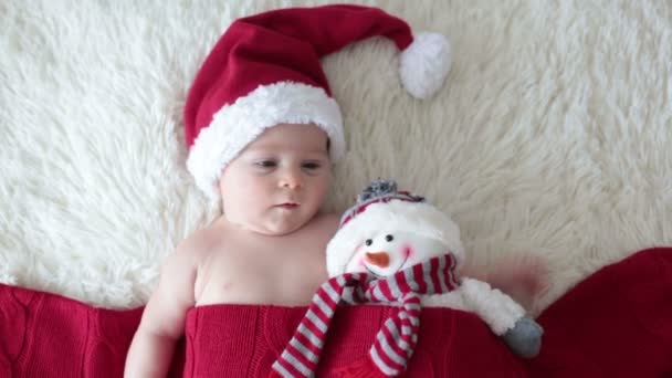 Christmas portrait of cute little newborn baby boy, wearing santa hat and hugging little cute snowman toy, studio shot, winter time - Кадри, відео