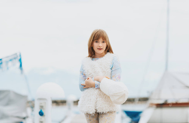 Portrait de mode en plein air de jeune fille préadolescente vêtue d'une casquette blanche, gilet en fausse fourrure, jouant dans un port, image tonique
 - Photo, image