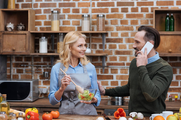 χαμογελαστή γυναίκα ανάμειξη σαλάτα στην κουζίνα και το σύζυγό της μιλάει με smartphone - Φωτογραφία, εικόνα