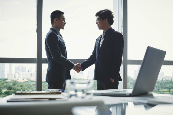 Профиль надежных деловых партнеров, пожимающих руки, стоя у панорамного окна просторного зала заседаний
 - Фото, изображение