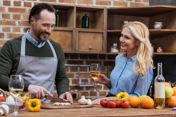 smiling husband cutting vegetables and wife standing with glass of wine - Foto, afbeelding