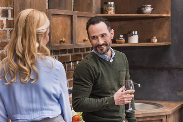 smiling husband with glass of wine talking to wife in kitchen - Zdjęcie, obraz