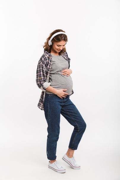 smiling pregnant woman listening music in headphones and touching belly isolated on white  - Valokuva, kuva