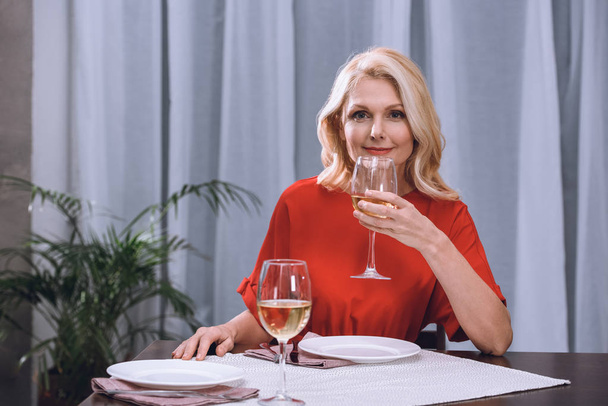 atraktivní žena v červených šatech pít víno u stolu - Fotografie, Obrázek