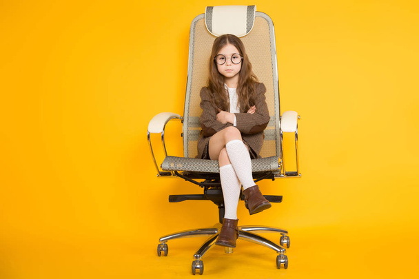 Schattig klein brunette meisje zittend in moderne bureaustoel op gele achtergrond  - Foto, afbeelding