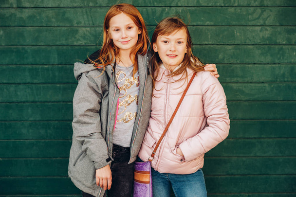Ulkona muotokuva kaksi söpöä pikku teini tytöt yllään lämpimät takit, poseeraa vihreää taustaa vasten, muoti lapsille
 - Valokuva, kuva
