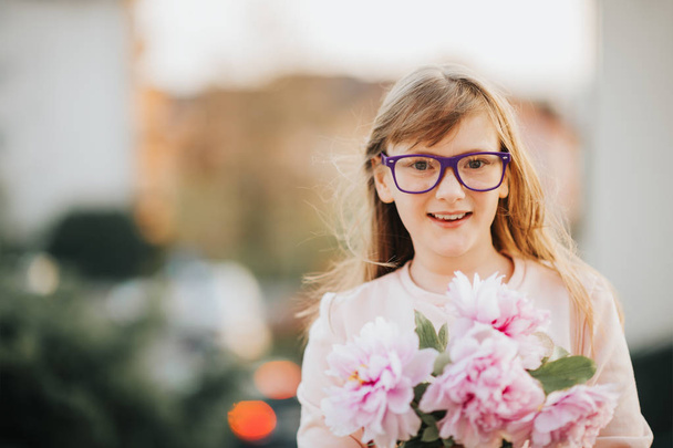 Outdoor portrait of a cute little 9 year old girl wearing eyeglasses, holding beautiful bouquet of fresh pink peonies - Photo, Image