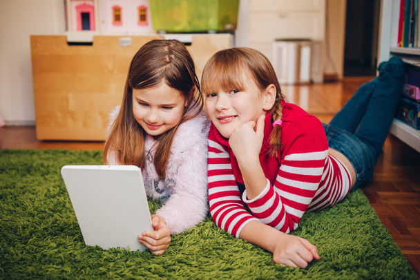 Two cute little girls playing on digital tablet pc, laying on green carpet in kid's room - Foto, imagen
