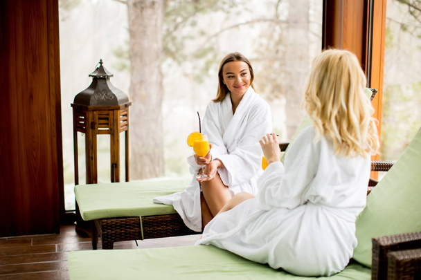 Two cheerful women in bathrobes drinking juice in spa center - Photo, Image