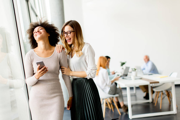 Jonge multiraciale zakenvrouwen glimlachend, terwijl andere ondernemers werken op achtergrond op kantoor - Foto, afbeelding