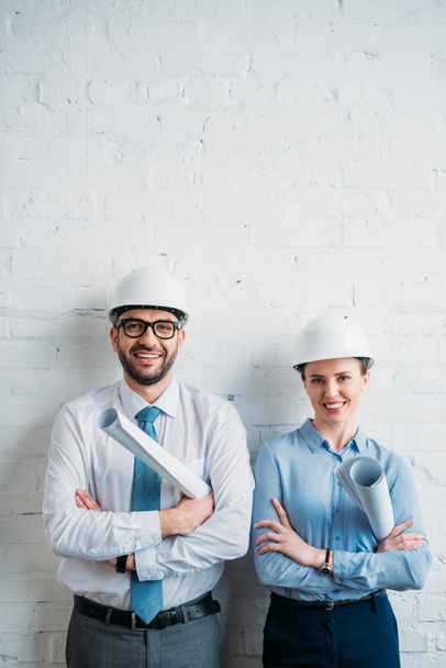 Lächelnde Architekten mit harten Hüten stehen vor weißen Backsteinwänden mit Bauplänen - Foto, Bild