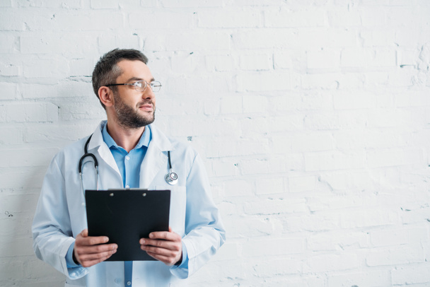 male doctor with clipboard in front of white brick wall - Photo, Image