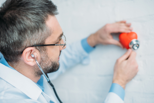 врач-мужчина слушает сердцебиение игрушечного сердца стетоскопами
 - Фото, изображение