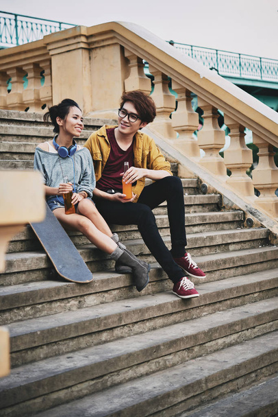 Cheerful Asian skateboarders sitting on shabby stairs, chatting and enjoying soft drinks, full length portrait - 写真・画像