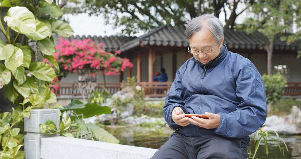 Азиатский старший мужчина смотрит по сотовому телефону в китайском саду
  - Фото, изображение