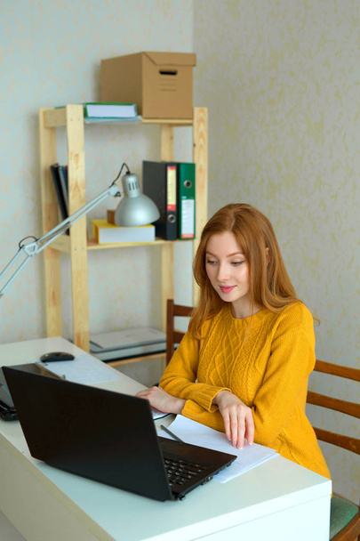 bela menina em uma camisola amarela e cabelo vermelho trabalhando em casa atrás de um laptop
 - Foto, Imagem
