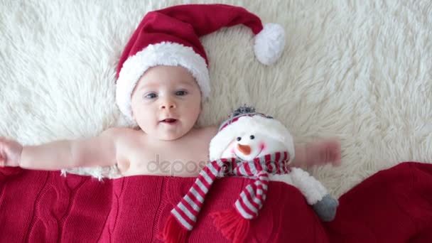 Christmas portrait of cute little newborn baby boy, wearing santa hat and hugging little cute snowman toy, studio shot, winter time - Кадри, відео