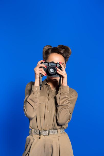 obscured view of fashionable young woman in retro clothing with photo camera isolated on blue - Foto, afbeelding