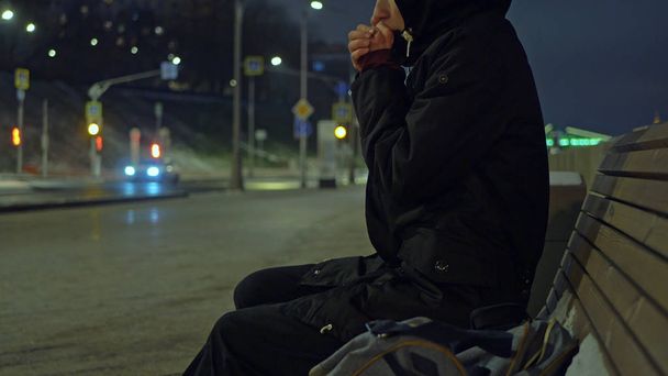 muž v kápi zahřeje na lavičce v noci na ulici během zasněžené zim - Fotografie, Obrázek