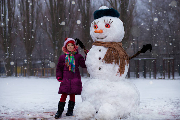 Een klein vrolijk meisje in de buurt van grote grappige sneeuwpop. Een schattig klein meisje heeft plezier in winter park, winter - Foto, afbeelding