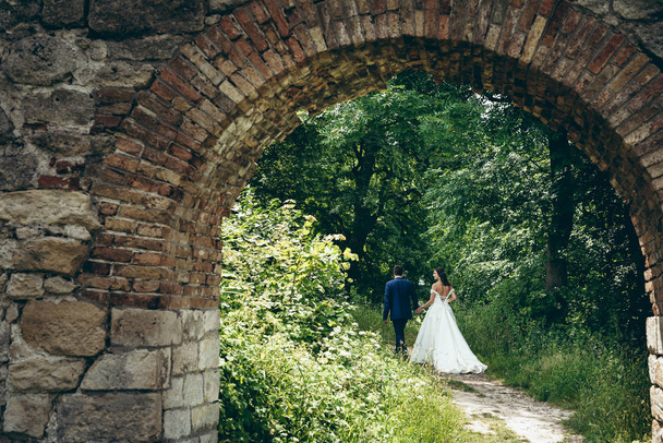 Rückansicht des schönen wandernden Brautpaares an der Hand entlang des Weges durch den grünen Wald. Blick durch den alten Steinbogen. - Foto, Bild