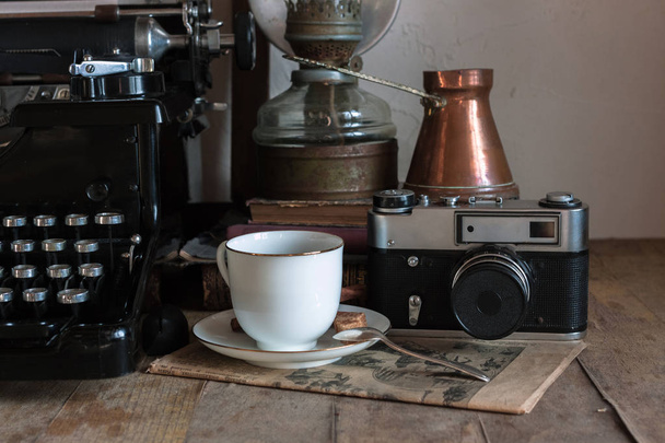 Λεπτομέρειες του still life στο εσωτερικό του σπιτιού. Vintage ρετρό δημοσιογράφος desktop με φλιτζάνι καφέ, χαλκό κανάτα, λαμπτήρας αερίου, βιβλία, εφημερίδα, typewrittert σε ξύλινο υπόβαθρο σε ρουστίκ στυλ. - Φωτογραφία, εικόνα