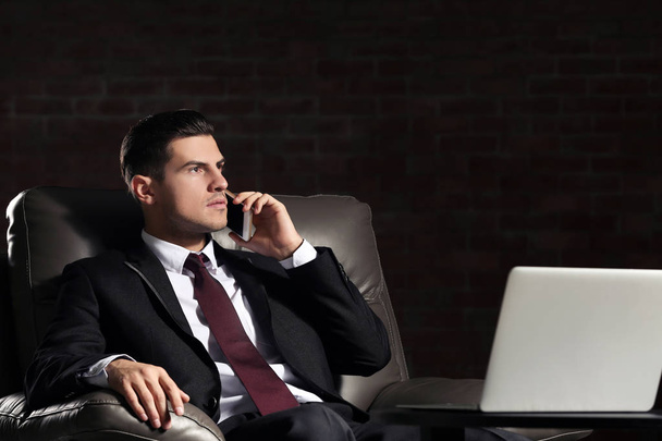 Handsome young man in suit talking by phone while sitting in armchair indoors - Photo, Image