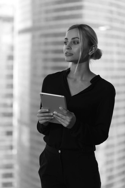 Hermosa mujer de negocios con tableta en las manos se encuentra cerca de la ventana en la oficina
. - Foto, Imagen