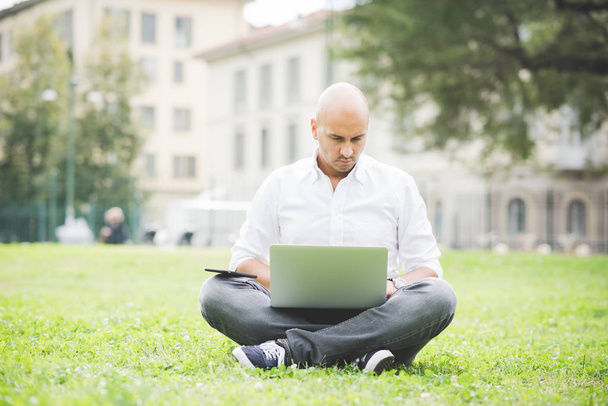Joven hombre de negocios contemporáneo trabajando a distancia sentado al aire libre en un parque de la ciudad usando smartphone y computadora
 - Foto, imagen