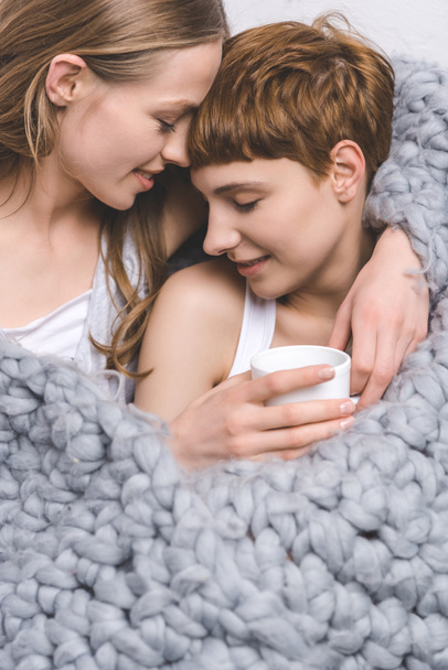 primer plano de feliz pareja lesbiana con taza de café abrazando debajo de punto manta de lana
 - Foto, Imagen