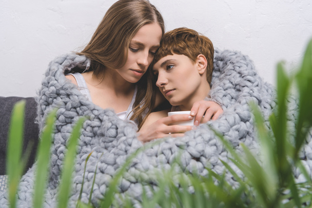 hermosa pareja lesbiana con taza de café abrazando debajo de la manta de lana de punto
 - Foto, Imagen