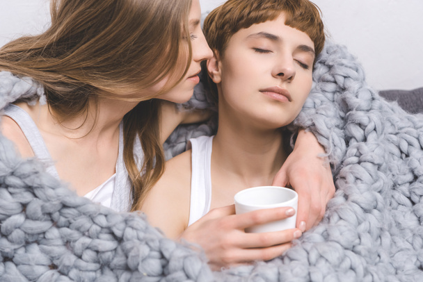 jovem casal lésbico com xícara de café abraçando sob cobertor de lã de malha
 - Foto, Imagem