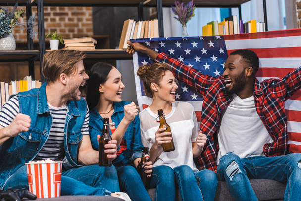 happy young multiethnic friends holding american flag and bottles of beer while sitting on sofa  - 写真・画像