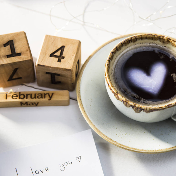The heart is reflected in a cup of coffee on the table next to the wooden calendar. Valentine's Day Concept - Photo, Image