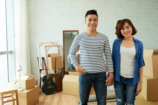 beautiful young Asian couple holding hands and standing in room with carton boxes - Фото, изображение