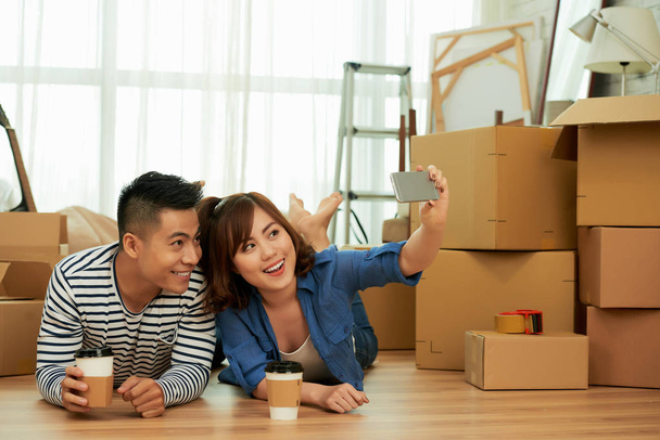 beautiful young Asian couple lying on floor in room with carton boxes and taking selfie photo on mobile phone - Photo, Image