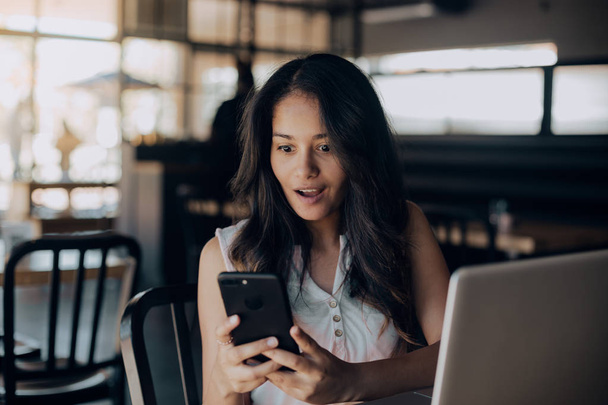 Vista frontal de la joven mestiza mirando su teléfono inteligente mientras está sentada en la cafetería
 - Foto, imagen