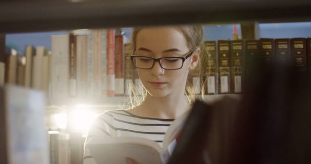 Portrait shot of the young female student in glasses reading a book among shelves with books in the library. Close up. Indoor - Фото, зображення