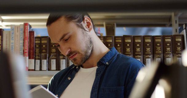 Portrait shot of the young male attractive student reading a book among shelves with books in the library. Close up. Inside - Фото, зображення