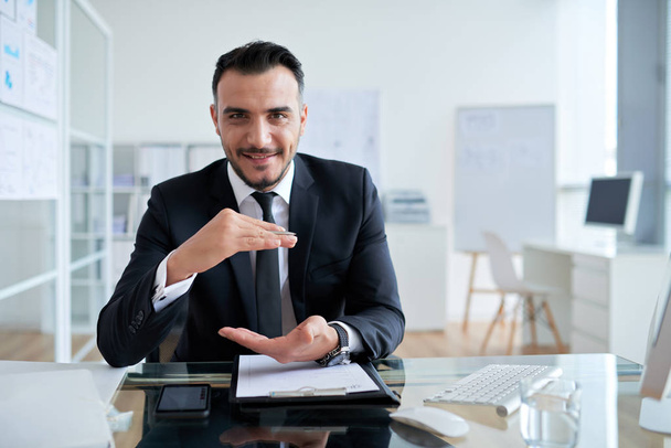Caucasian businessman gesturing with hands while sitting at workplace table in office and looking at camera  - Photo, image