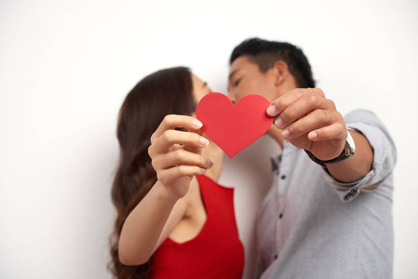 Perfil vista de afetuoso casal asiático beijando enquanto em pé contra o fundo branco e mostrando cartão de São Valentim para câmera, estúdio tiro
 - Foto, Imagem