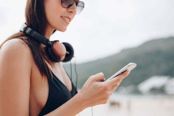 mulher atraente em óculos de sol e boné com fones de ouvido usando smartphone
 - Foto, Imagem