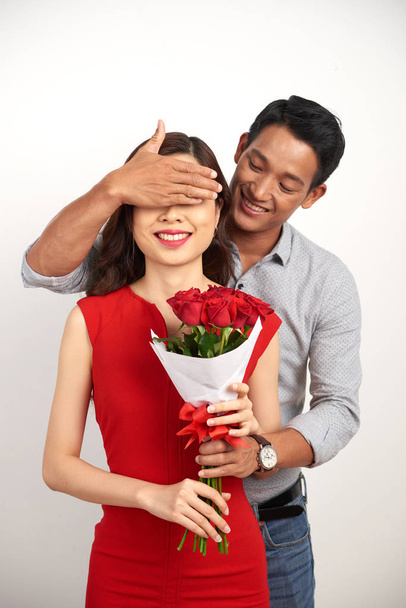 Handsome Asian man covering eyes of his smiling girlfriend with hand while passing her bouquet of red roses, isolated white background - Foto, immagini